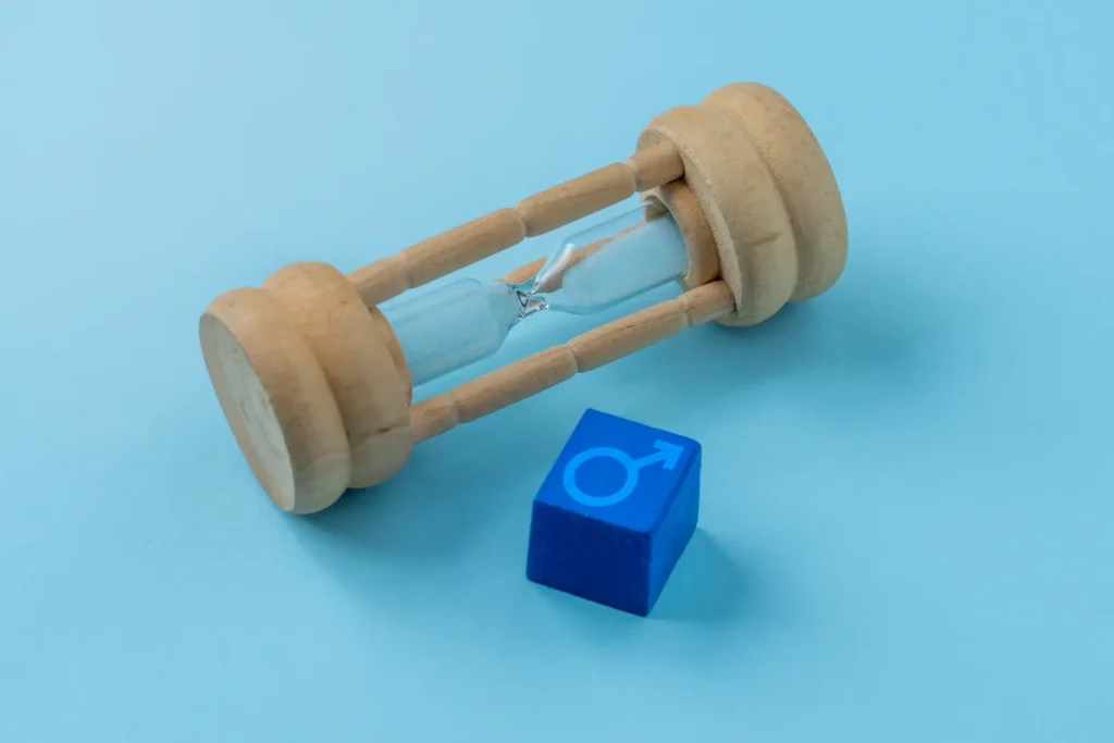 Wooden hourglass beside a blue block marked with a male symbol on a light blue surface.